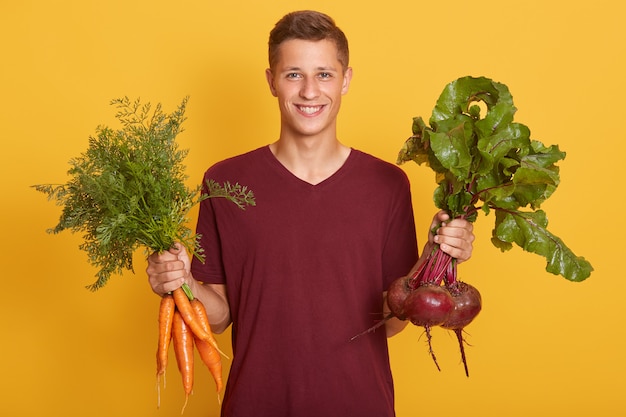 Glücklicher fröhlicher junger blonder Mann, der gesundes Gemüse für Salat hält.