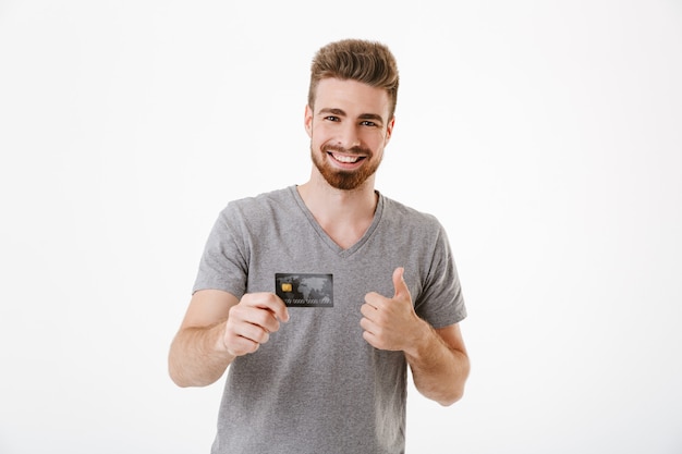 Glücklicher fröhlicher junger Mann, der Kreditkarte hält, macht Daumen hoch Geste.