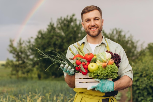 Glücklicher Gärtnermann, der Korb mit frischem Gemüse auf Regenbogen- und Gartenhintergrund-Gartenkonzept hält