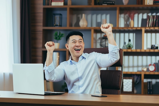 Glücklicher Geschäftsmann, der an einem Tisch in einem klassischen asiatischen Büro tanzt und den Sieg feiert, der am Computer arbeitet