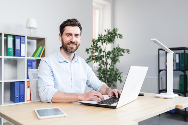 Glücklicher Geschäftsmann, der in einem hellen, modernen Büro hinter einem Laptop-Mann mit Bart arbeitet, lächelt und in die Kamera schaut