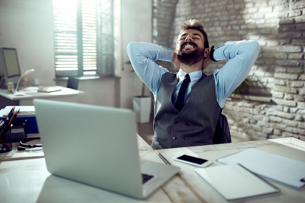 Glücklicher Geschäftsmann, der sich mit den Händen hinter dem Kopf entspannt, während er eine Pause von der Arbeit im Büro macht