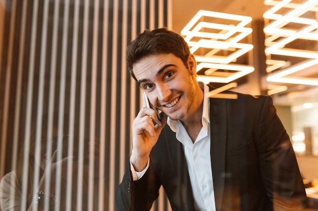 Glücklicher Geschäftsmann im Anzug, der im Büro hinter dem Glas telefoniert