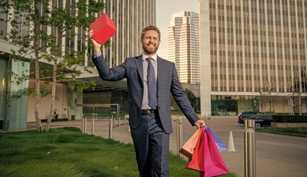 Glücklicher Geschäftsmann im Anzug hält Pakete und Geschenkboxen, die vor dem Bürogruß spazieren gehen