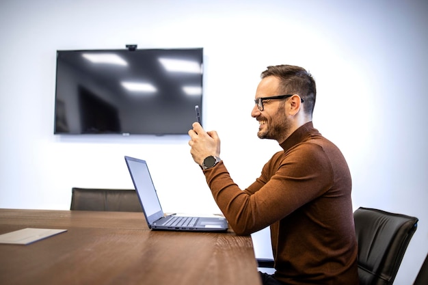 Glücklicher Geschäftsmann in Freizeitkleidung, der sein Smartphone nutzt, während er in seinem Büro arbeitet