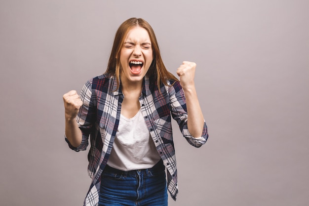 Glücklicher Gewinner! Nahaufnahme der emotionalen jungen attraktiven Frau mit Händchenhalten in den Fäusten, lokalisiert auf grauer Wand. Überraschte junge Frau schreit.