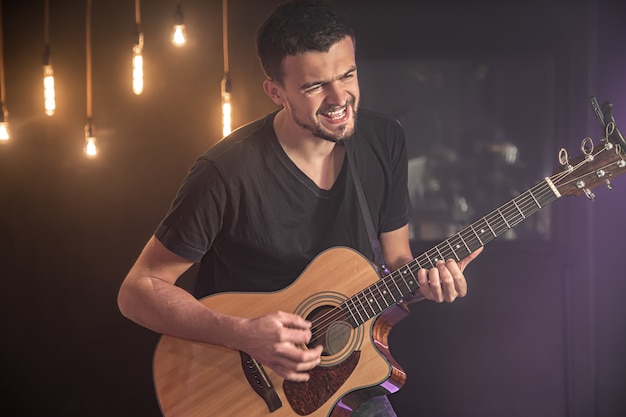 Glücklicher Gitarrist in einem schwarzen T-Shirt spielt eine Akustikgitarre bei einem Konzert vor einem verschwommenen schwarzen Hintergrund.