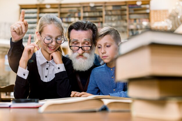 Glücklicher Großvater, der Buch mit Enkel und Enkelin liest und am Tisch in der alten Weinlesebibliothek sitzt. Der ältere Mann und das Mädchen zeigen mit den Fingern nach oben und lachen