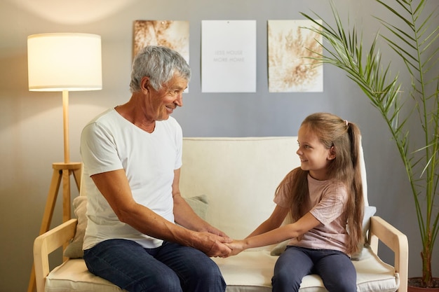 Glücklicher Großvater und seine Enkelin sitzen zusammen auf dem Sofa zu Hause im Wohnzimmer und unterhalten sich Händchen haltend über die Probleme ihrer Kinder