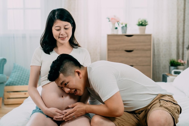 glücklicher gutaussehender asiatischer chinesischer mann, der dem bauch der schönen schwangeren frau zuhört. Geschenk für ungeborenes Baby für Papa am Vatertag. charmante werdende mutter, die ihren fröhlichen ehemann mit nacktem bauch anschaut, sitzt auf dem bett.