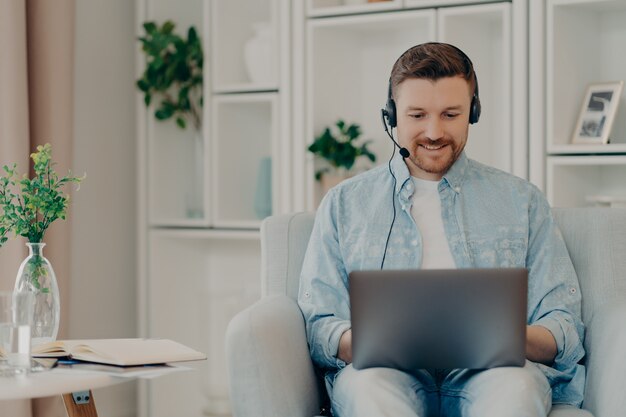 Glücklicher gutaussehender Mann in Freizeitkleidung, der online chattet und Headset und Laptop verwendet, während er zu Hause in einem bequemen Sessel im Wohnzimmer sitzt. Freiberufler mit Videokonferenz am Computer