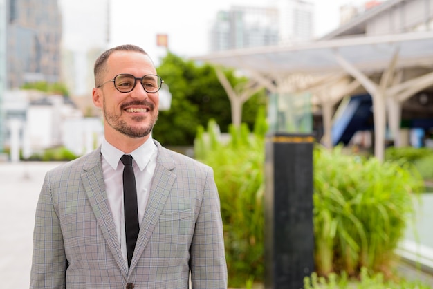 Glücklicher hispanischer kahler bärtiger Geschäftsmann mit Brillen, die in der Stadt draußen denken