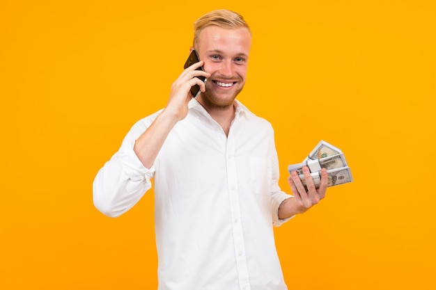 Glücklicher hübscher blonder Mann in einem weißen Hemd hält ein Bündel Papiergeldscheine und spricht am Telefon auf Gelb