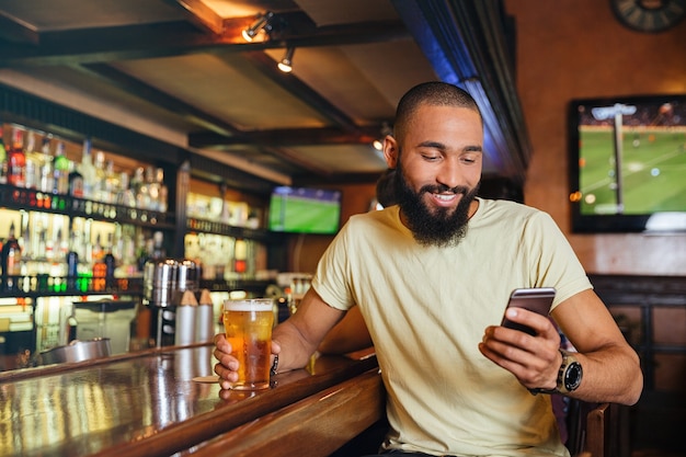 Glücklicher hübscher junger Mann, der Bier in der Bar trinkt und Smartphone benutzt