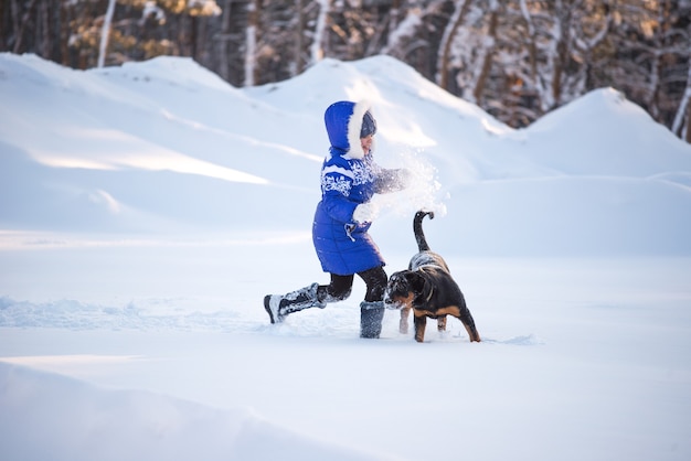 Glücklicher Hund und ein freudiges Mädchen, das auf einem verschneiten Feld nahe dem Wald an einem sonnigen Wintertag läuft.