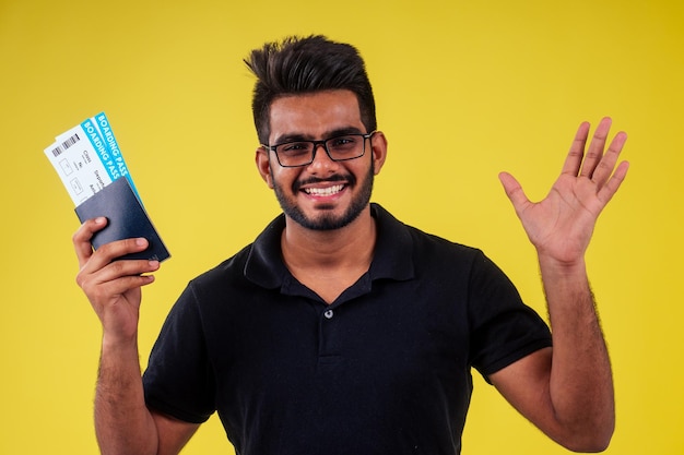 Glücklicher indischer bärtiger Student mit Brille und schwarzem Tshort mit Passflugticket gelbem Studiohintergrund Lifestyle-Sommerreisekonzept
