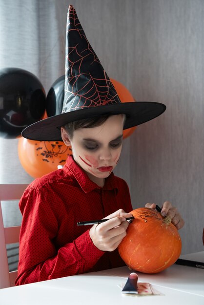 Glücklicher jugendlich Junge im Kostüm, das sich auf die Halloween-Feier vorbereitet, die einen Kürbis zeichnet