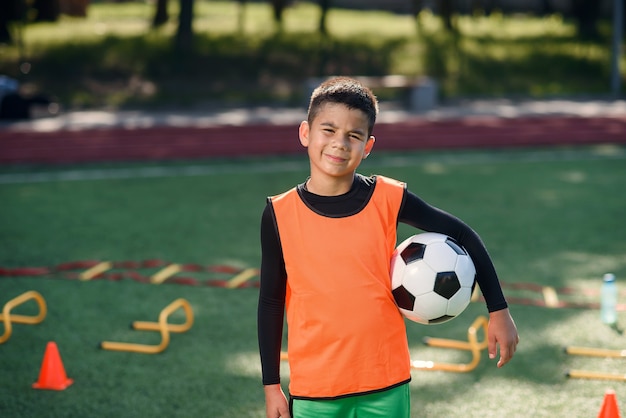 Glücklicher jugendlich Junge in der Fußballuniform hält einen Ball nach dem Morgentraining auf dem Stadion