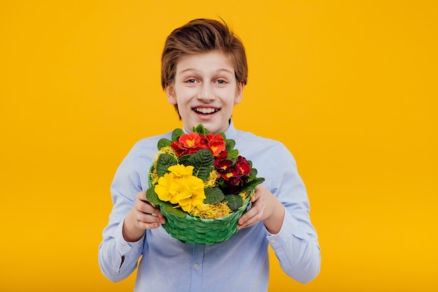 Glücklicher jugendlicher junge mit blumenkorb in seiner hand, im blauen hemd, lokalisiert auf gelber wand