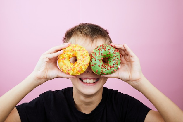 Glücklicher Junge hat Spaß, der mit Donuts auf rosa Hintergrund gespielt wird. Kind hält Donutgläser.