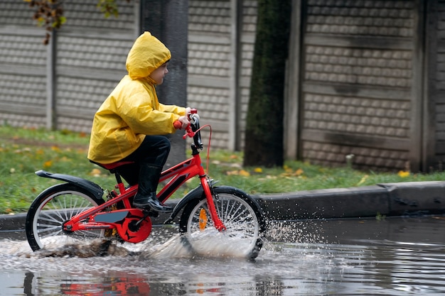 Glücklicher Junge im Regenmantel macht Spaß mit dem Fahrrad durch die Pfützen. Kind auf Fahrrad im Regen.