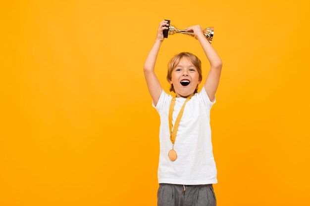 Glücklicher Junge in einem T-Shirt mit einer Medaille am Hals hebt den Siegerpokal auf Gelb mit Kopierraum an