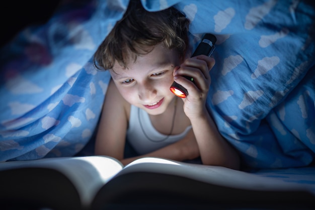 Glücklicher Junge liegt nachts unter Decke und liest Buch mit Taschenlampe