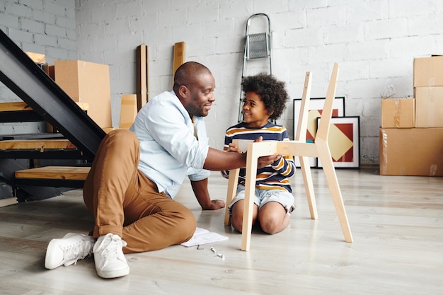 Glücklicher junger Afrikaner, der seinen süßen kleinen Sohn anschaut, während er ihm beibringt, wie man einen Holzstuhl zusammenbaut, während er beide auf dem Boden sitzt