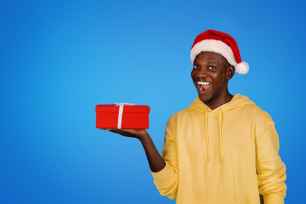 Glücklicher junger afrikanisch-amerikanischer Kerl in Sweatshirt und Weihnachtsmütze zeigt Geschenkbox