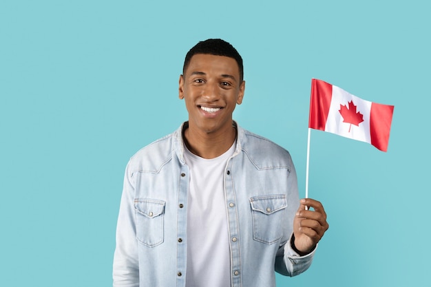 Glücklicher junger afrikanisch-amerikanischer männlicher Student in ungezwungener Show Kanada-Flagge isoliert auf blauem Hintergrund Studio