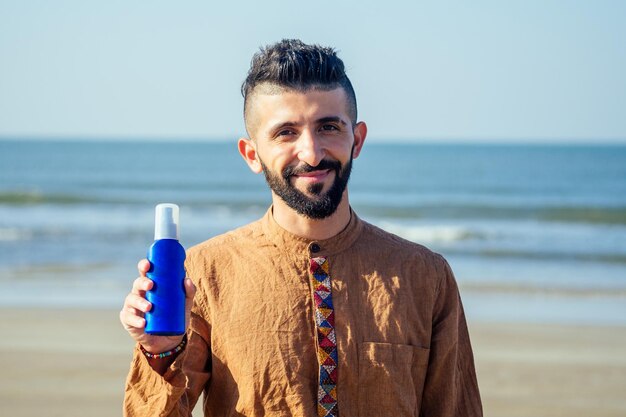 Glücklicher junger arabischer Mann, der Sonnencreme verwendet, schützt sich am StrandIndischer Mann, der eine Flasche Sonnencreme hält