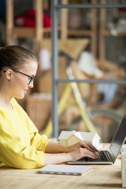 Glücklicher junger Architekt oder Ingenieur im lässigen gelben Pullover, der durch Tisch vor Laptop sitzt und im Netz nach Daten sucht