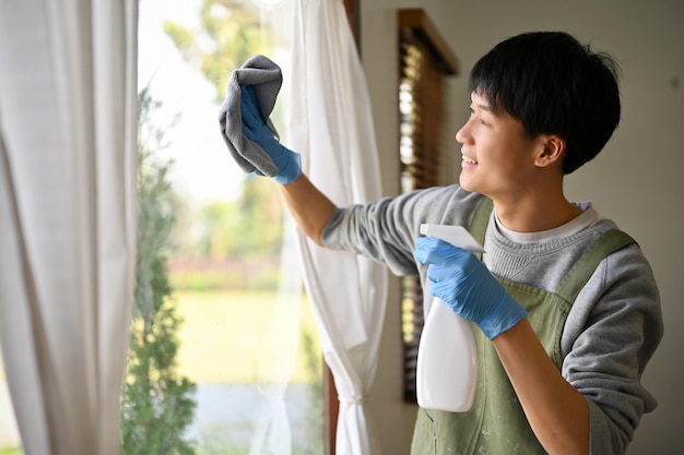 Glücklicher junger asiatischer Mann, der Glasspray und Staubtuch verwendet, um das Fensterglas zu reinigen