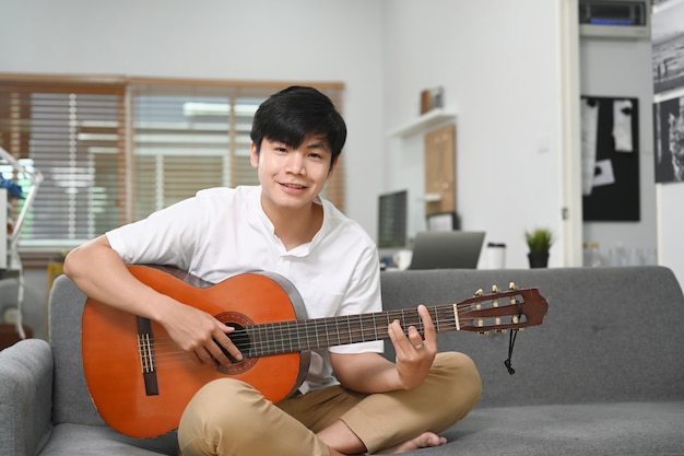 Glücklicher junger asiatischer Mann, der zu Hause mit seiner Gitarre auf dem Sofa lächelt und sitzt.