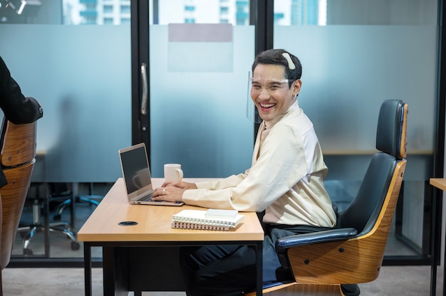 Glücklicher junger asiatischer Transvestit-Büroangestellter, der mit Laptop und Dokument am Schreibtisch im Büro arbeitet