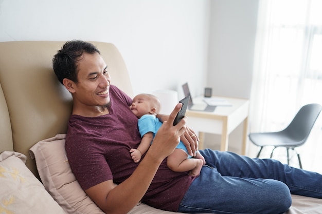 Glücklicher junger asiatischer Vater, der sein neugeborenes süßes entzückendes Baby hält, das auf seinen Armen schläft, während Handy auf dem Bett benutzt wird