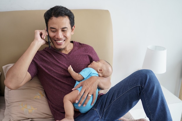 Glücklicher junger asiatischer Vater, der sein neugeborenes süßes entzückendes Baby hält, das auf seinen Armen schläft, während Handy auf dem Bett benutzt wird