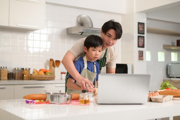 Glücklicher junger asiatischer Vater und Kind nutzen einen Laptop, um zu lernen, wie man Essen kocht