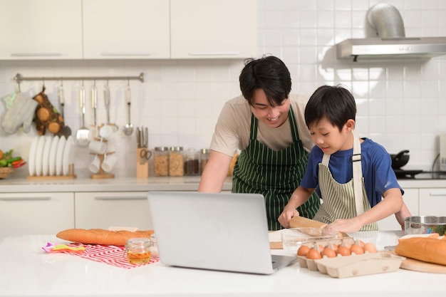 Glücklicher junger asiatischer Vater und Kind nutzen einen Laptop, um zu lernen, wie man Essen kocht