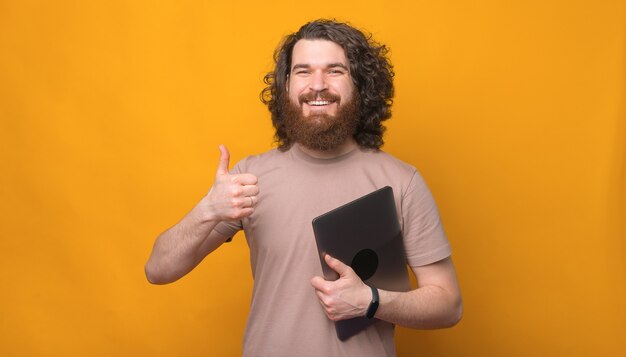 Glücklicher junger bärtiger Mann lächelnd, der Laptop hält und Daumen oben Geste über Gelb zeigt
