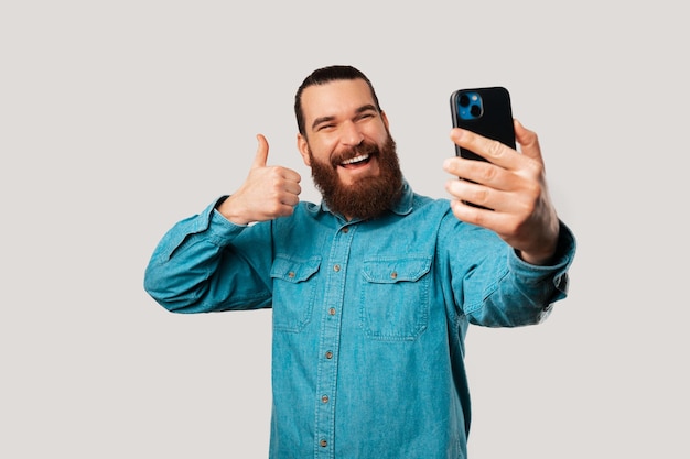 Glücklicher junger bärtiger Mann macht ein Selfie mit seinem Telefon und zeigt einen Daumen nach oben