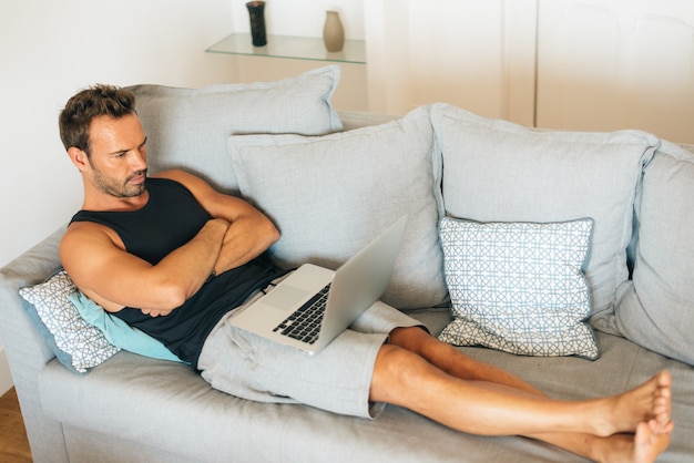 Glücklicher junger blonder Mann, der sich zu Hause in der Couch auf dem Computer hinlegt