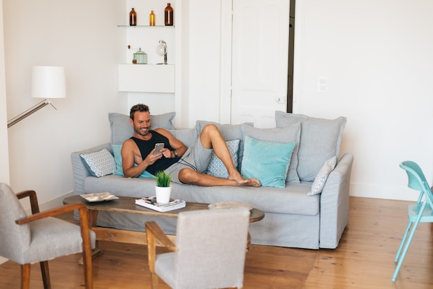 Glücklicher junger blonder Mann, der zu Hause in der Couch am Handy sitzt