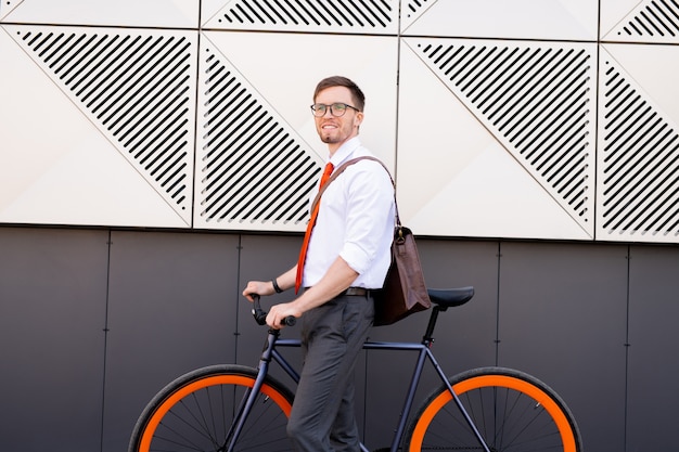 Glücklicher junger eleganter Mann, der an der Wand der modernen Architektur steht, während er Fahrrad benutzt, um nach Arbeitstag nach Hause oder zum Café zu gehen