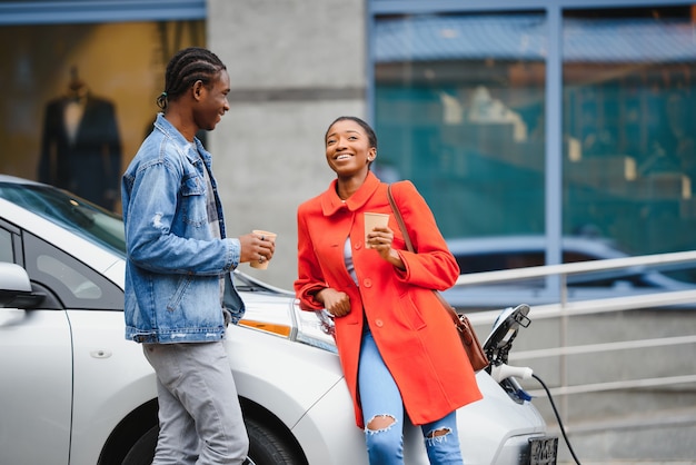 Glücklicher junger erwachsener Afroamerikanermann und lächelnde Frau, die Elektroauto auflädt.