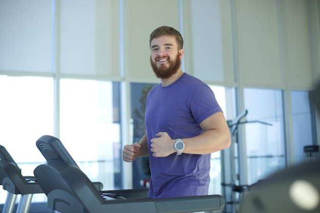 Glücklicher junger, fitter Mann, der im Fitnessstudio trainiert, joggt auf dem Laufband, Cardio-Training macht und lächelt