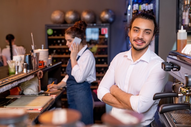 Glücklicher junger gekreuzter Kellner im weißen Hemd, das vor Kamera auf Hintergrund des Kollegen steht, der mit Kunden am Telefon spricht