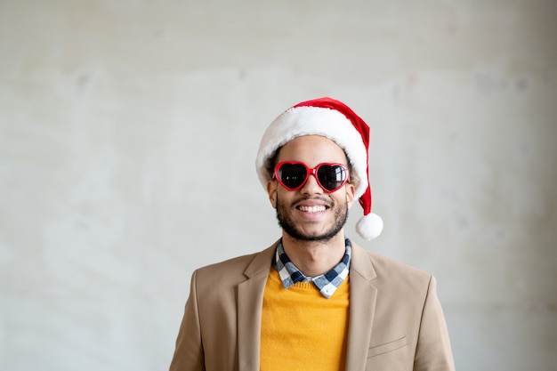 Glücklicher junger Geschäftsmann mit gemischten Rassen in eleganter Freizeitkleidung, Weihnachtsmütze und herzförmiger Sonnenbrille, der vor der Kamera lächelt