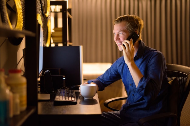 Glücklicher junger Geschäftsmann mit Kaffee, der am Telefon spricht, während er spät abends von zu Hause aus arbeitet