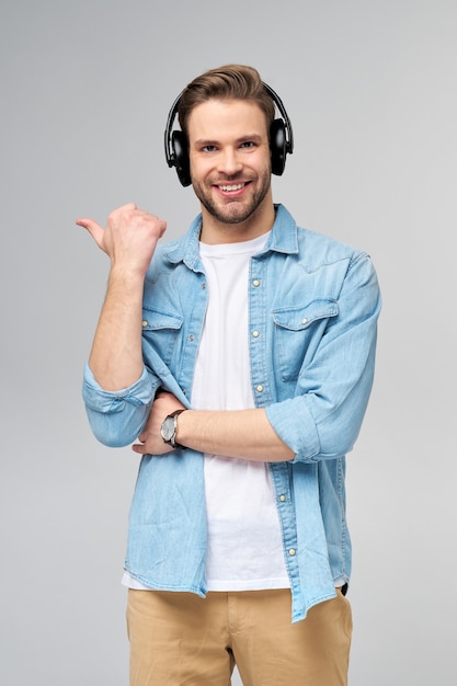 Glücklicher junger gutaussehender Mann im Jeanshemd, der weg zeigt gegen graue Wand, die große Kopfhörer trägt